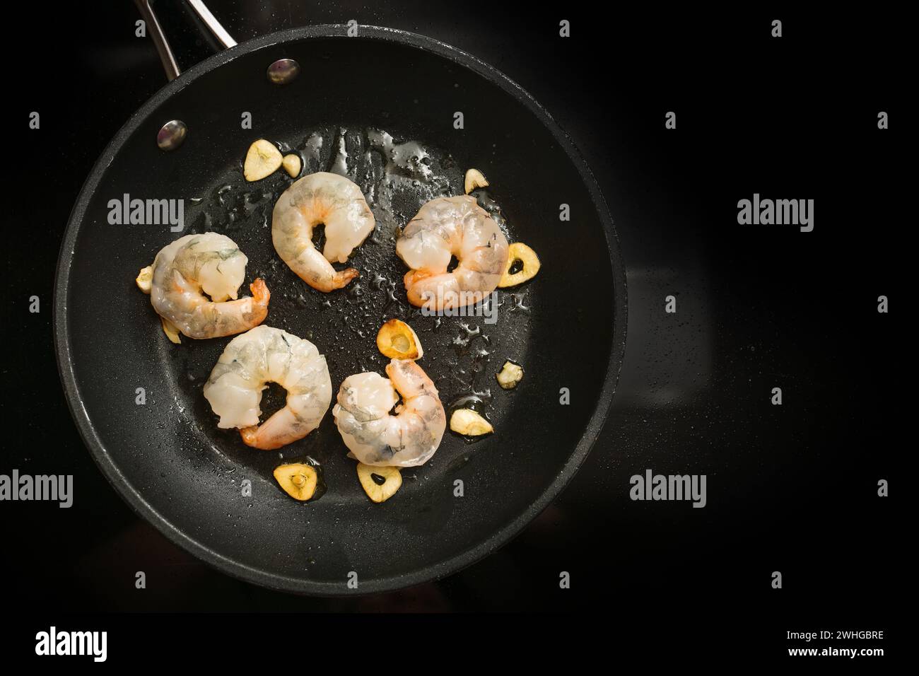 I gamberetti freschi vengono saltati con aglio in olio d'oliva in una pentola sul piano cottura nero, per preparare un delizioso antipasto, c Foto Stock