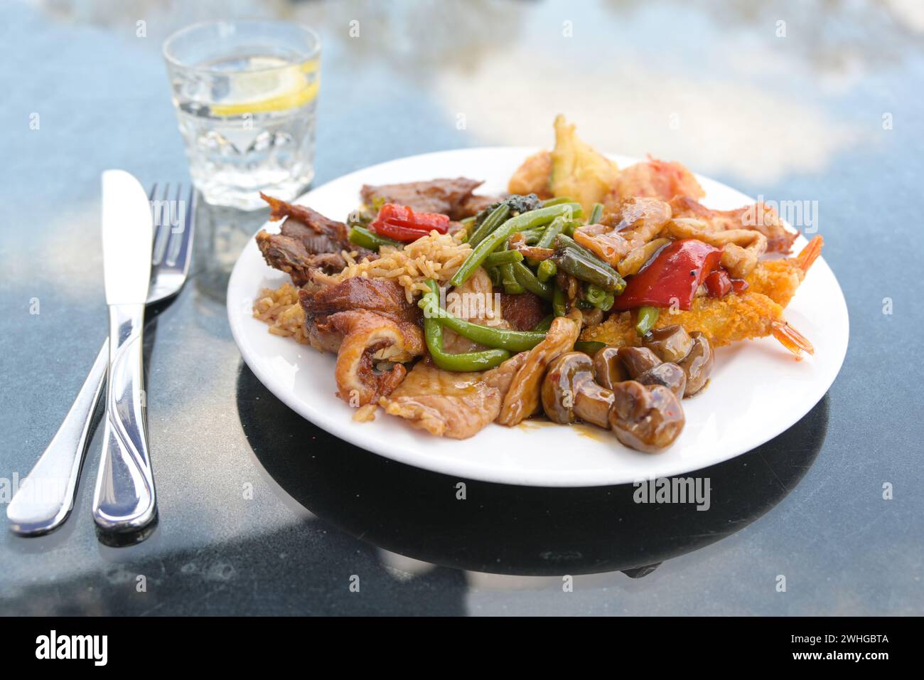 Piatto cinese in un ristorante di strada, vari tipi di carne e verdure con riso fritto su un piatto bianco, posate e un bicchiere con wate Foto Stock