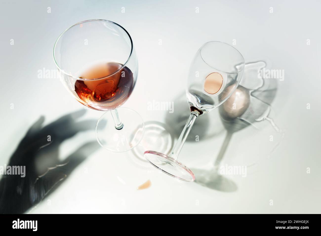 Bicchieri con vino rosso, in piedi e caduti in una prospettiva insolita con riflessi e un'ombra di mano su uno sfondo luminoso Foto Stock