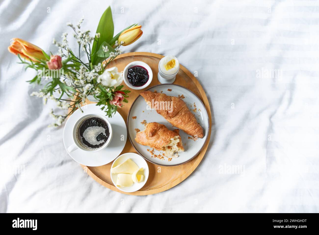 Colazione tranquilla a letto, vassoio di legno con croissant, caffè, marmellata, uova, burro e fiori su biancheria bianca verde grigia, domenica o. Foto Stock