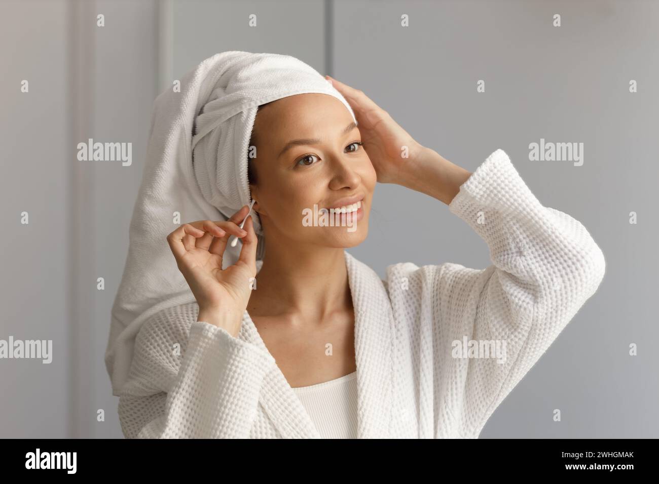 La bella donna in abito bianco e con un asciugamano sulla testa pulisce le orecchie con bastoncino di cotone. Pulizia e cura sana del corpo. Portra Foto Stock