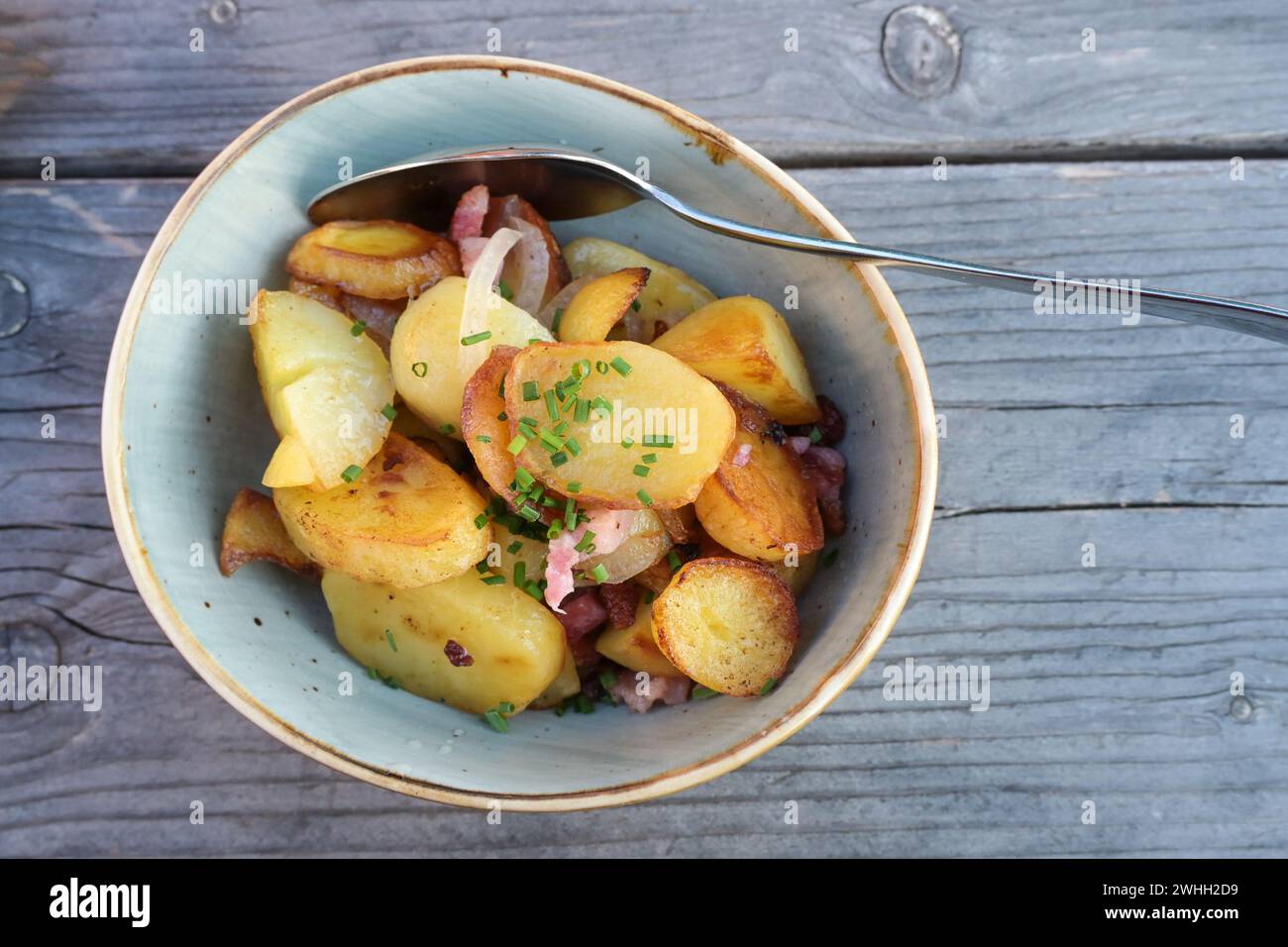 Patate fritte con cipolle, pancetta e cipollina guarnire in un recipiente di ceramica su un tavolo di legno grigio blu, vista dall'alto, c Foto Stock
