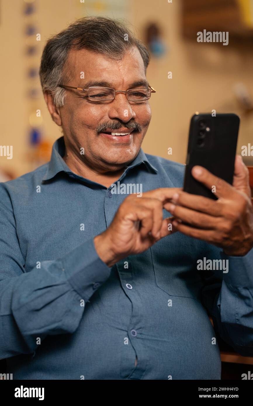 Scatto verticale, uomo anziano sorridente e felice che utilizza il telefono cellulare con occhiali da vista a casa - concetto di stile di vita pensionistico, contenuti sui social media e cyberspazio Foto Stock
