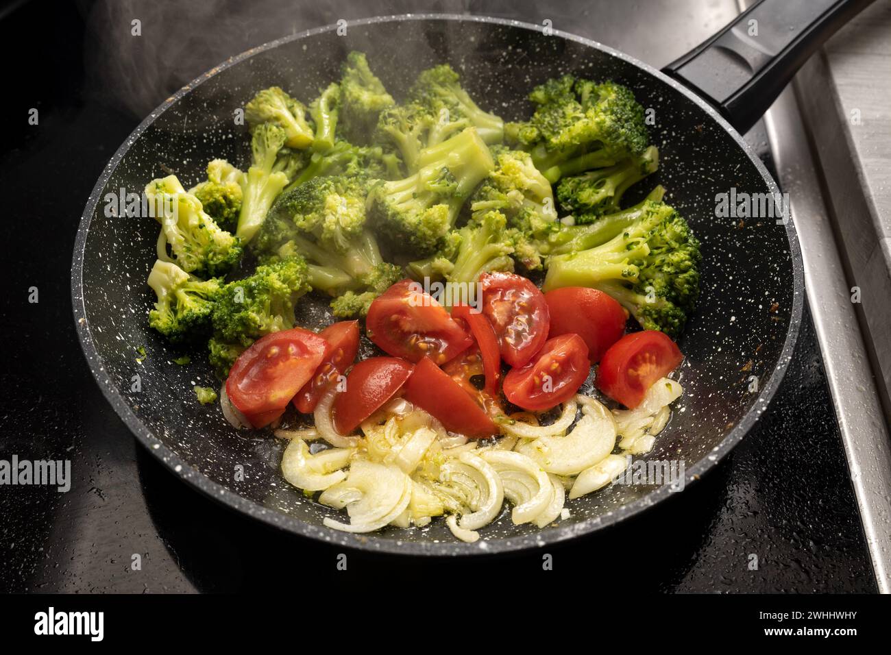 Cipolle, pomodori e broccoli vengono saltati in una padella al vapore sul piano cottura nero per un sano pasto vegetariano, selezionato Foto Stock