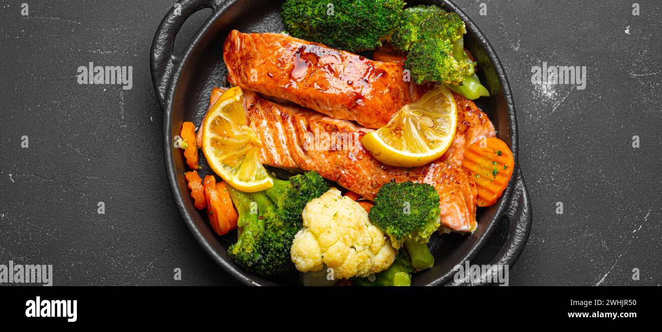 Pesce sano al forno bistecche di salmone, broccoli, cavolfiore, carota in casseruola in ghisa su fondo di pietra nera scura. Cuoco Foto Stock