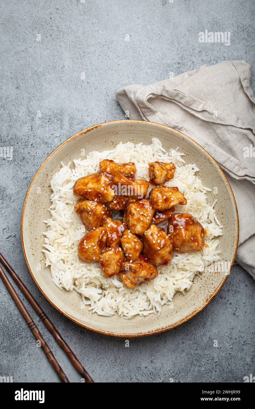 Pollo cinese agrodolce con semi di sesamo e riso su piatto di ceramica con bacchette vista dall'alto, pietra rustica grigia ba Foto Stock