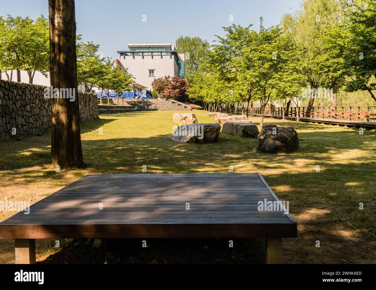 Tavolo da picnic in legno in un parco pubblico ombreggiato da alberi con una passerella in legno sul lato destro della cornice e un muro in pietra sul lato sinistro della cornice Foto Stock