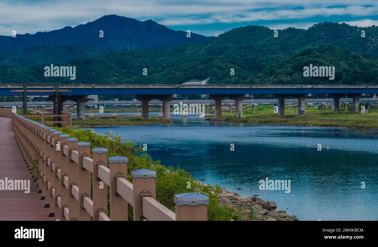 Marciapiede in legno lungo una strada di campagna accanto al fiume con ponte autostradale e montagne sotto il cielo nuvoloso sullo sfondo della Corea del Sud Foto Stock