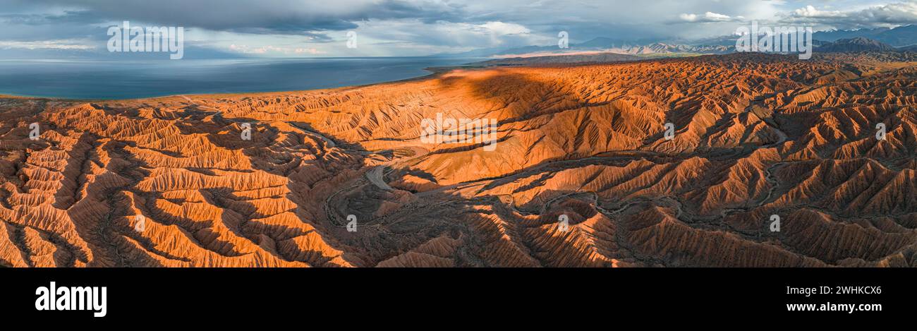 Panorama, paesaggio di colline erose, calanchi al tramonto, dietro il lago Issyk Kul e le vette delle montagne Tien Shan, vista aerea, Canyon of Foto Stock