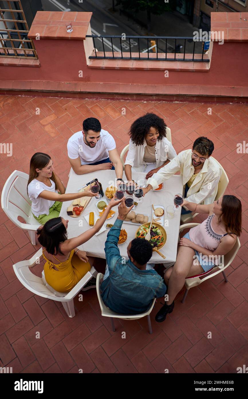 Dall'alto foto di giovani amici diversi che brindano bicchieri di vino rosso per celebrare il pranzo sul tetto. Foto Stock
