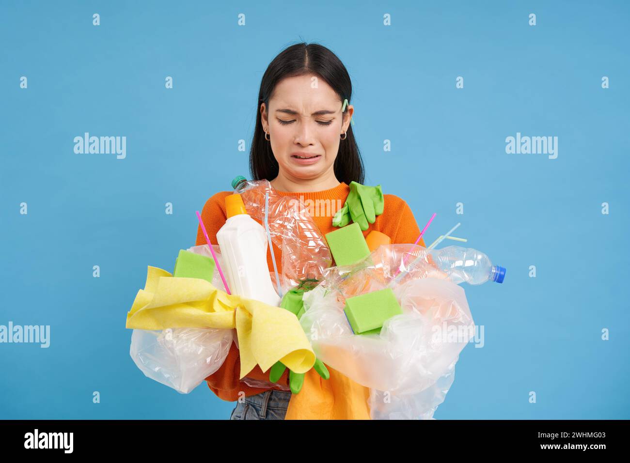 Donna asiatica scioccante con spazzatura di plastica in mano, lamentandosi di rifiuti non riciclabili, in piedi con i rifiuti sopra il backgroun blu Foto Stock