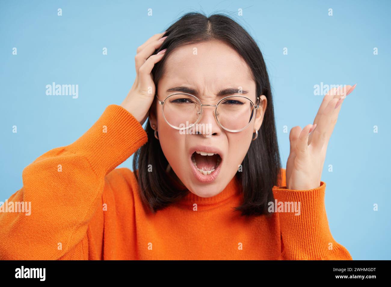 Ritratto di una giovane donna asiatica in occhiali, che litiga, urla e sembra infastidita, in piedi su sfondo blu Foto Stock