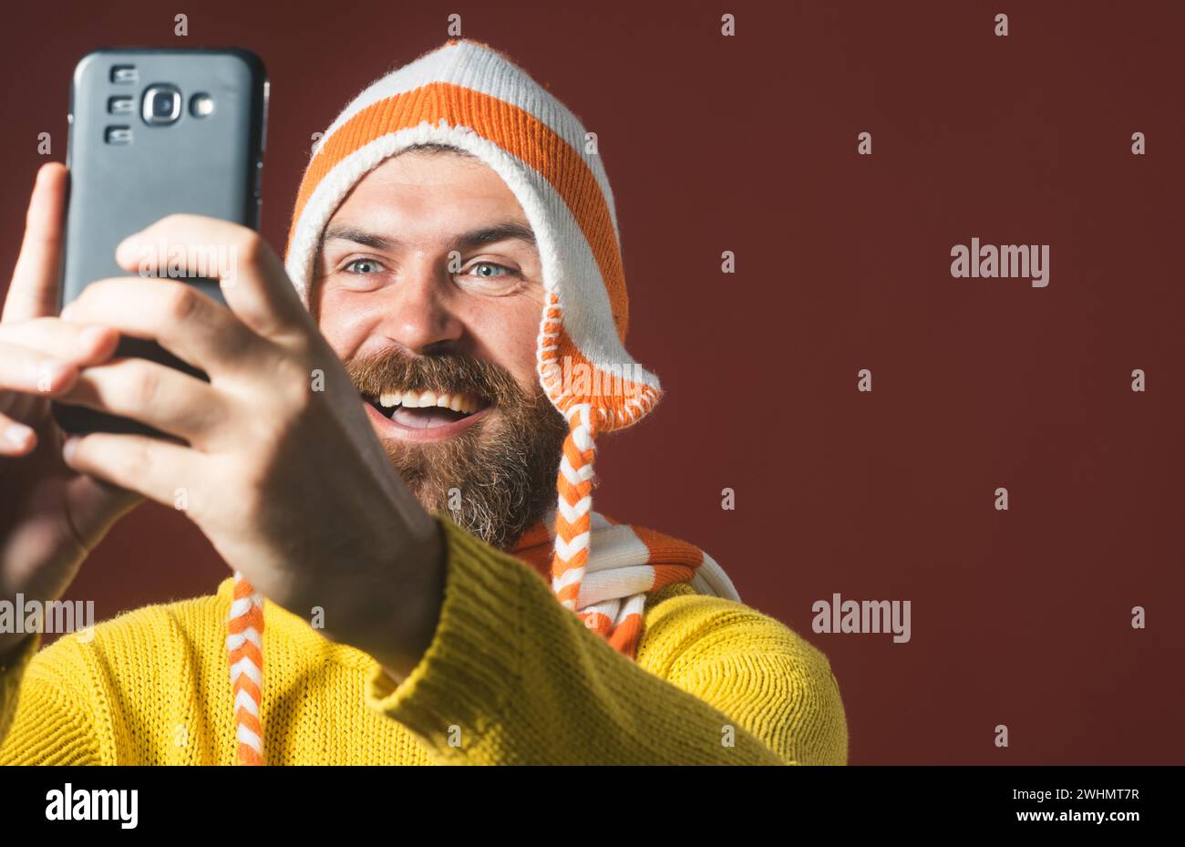 Tempo di selfie. Uomo sorridente che fa autoritratti sullo smartphone. Bell'uomo in maglione giallo fa selfie. Uomo barbuto felice con cappello divertente Foto Stock