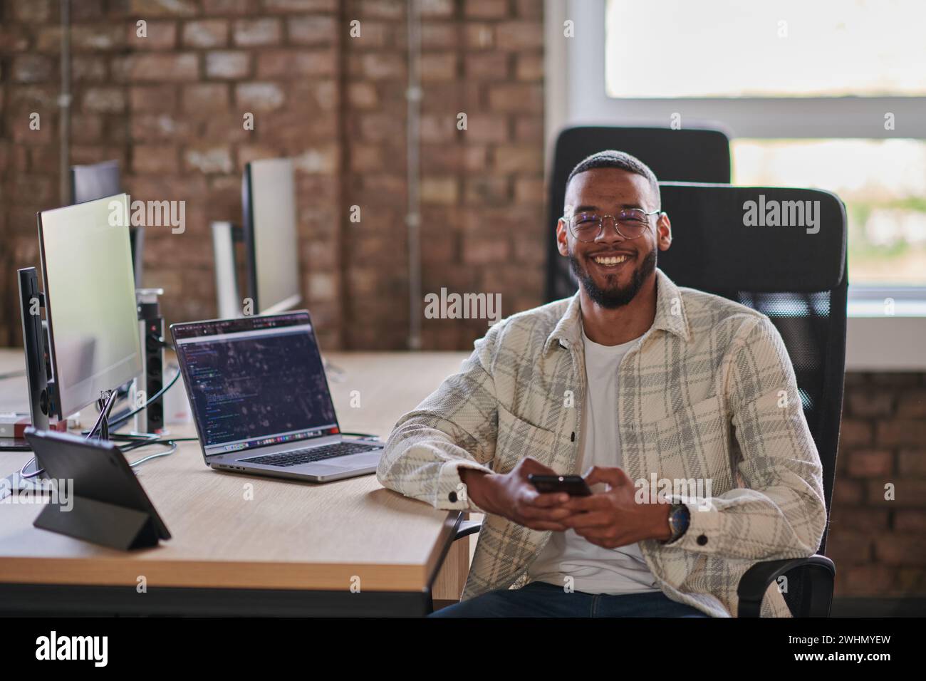 Un giovane imprenditore afroamericano si prende una pausa in un ufficio moderno, utilizzando uno smartphone per navigare sui social media, catturando un m Foto Stock