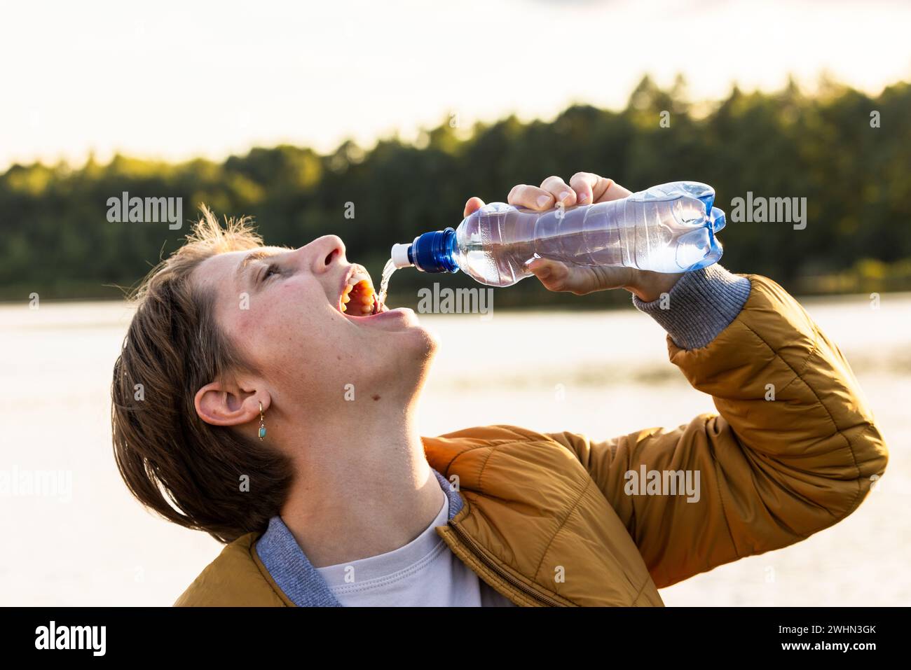 La giovane ragazza caucasica beve acqua rinfrescante da una bottiglia all'aperto. La bruna a occhi aperti indossa una camicia bianca e marrone Foto Stock
