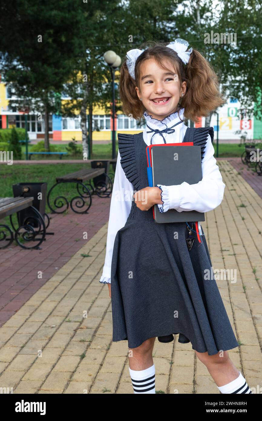 Ragazza con zaino, uniforme scolastica con archi bianchi e pila di libri vicino a scuola. Tornate a scuola, allievo felice, libri di testo pesanti. Foto Stock