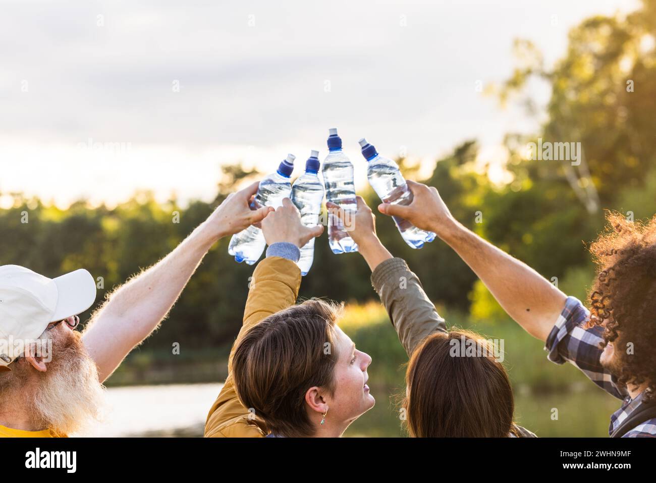 Gruppo multirazziale di amici che festeggiano e festeggiano fuori in natura tenendo in aria le loro bottiglie d'acqua di plastica Foto Stock