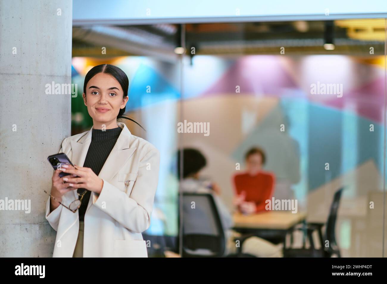 In un moderno ufficio in vetro, una donna d'affari contemporanea può operare in multitasking senza sforzo, utilizzando il suo smartphone, mentre è circondata da collea Foto Stock
