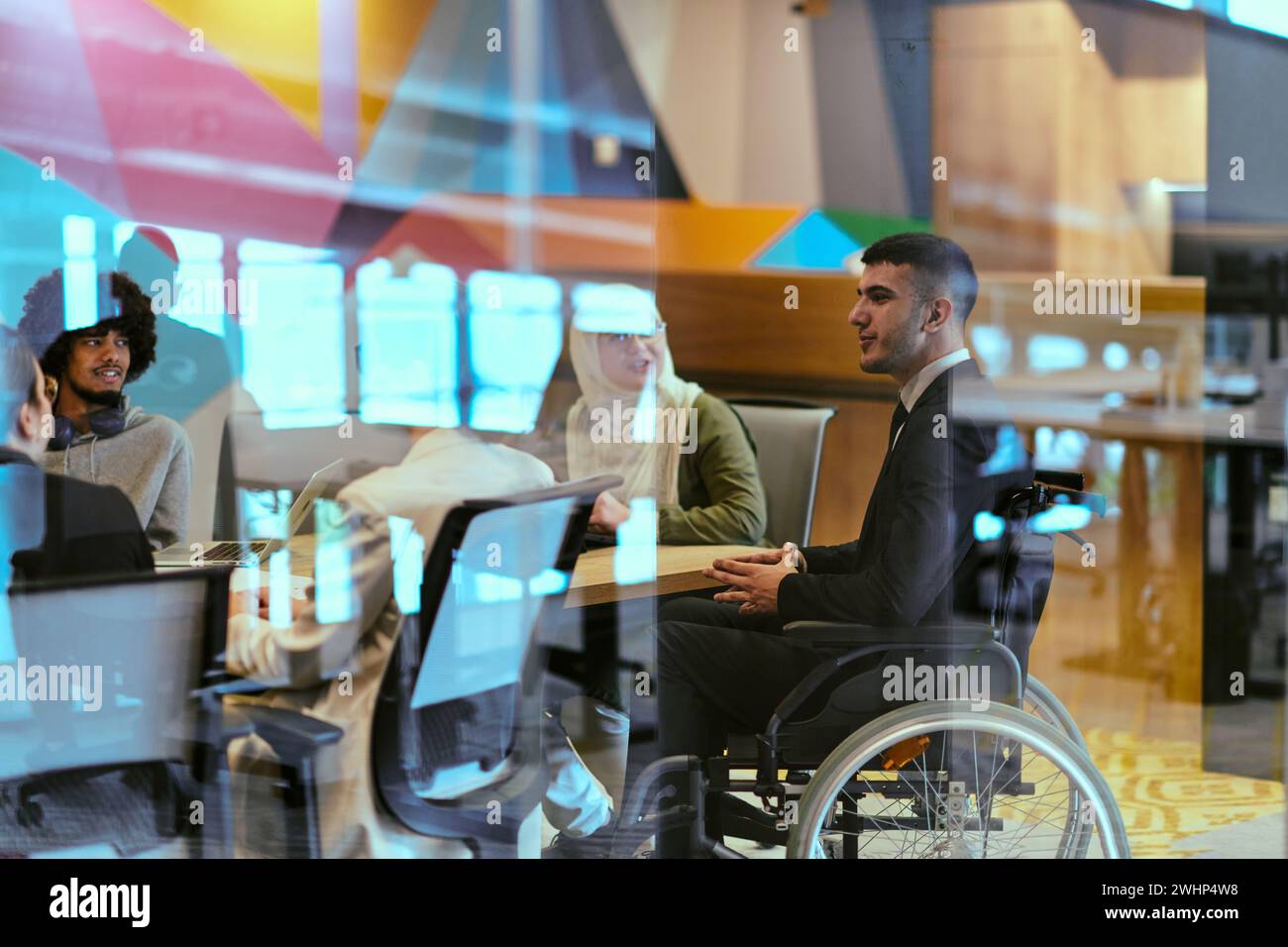 In un moderno ufficio di startup in vetro, un direttore in sedia a rotelle conduce un incontro di successo con i colleghi, incarnando l'inclusività Foto Stock