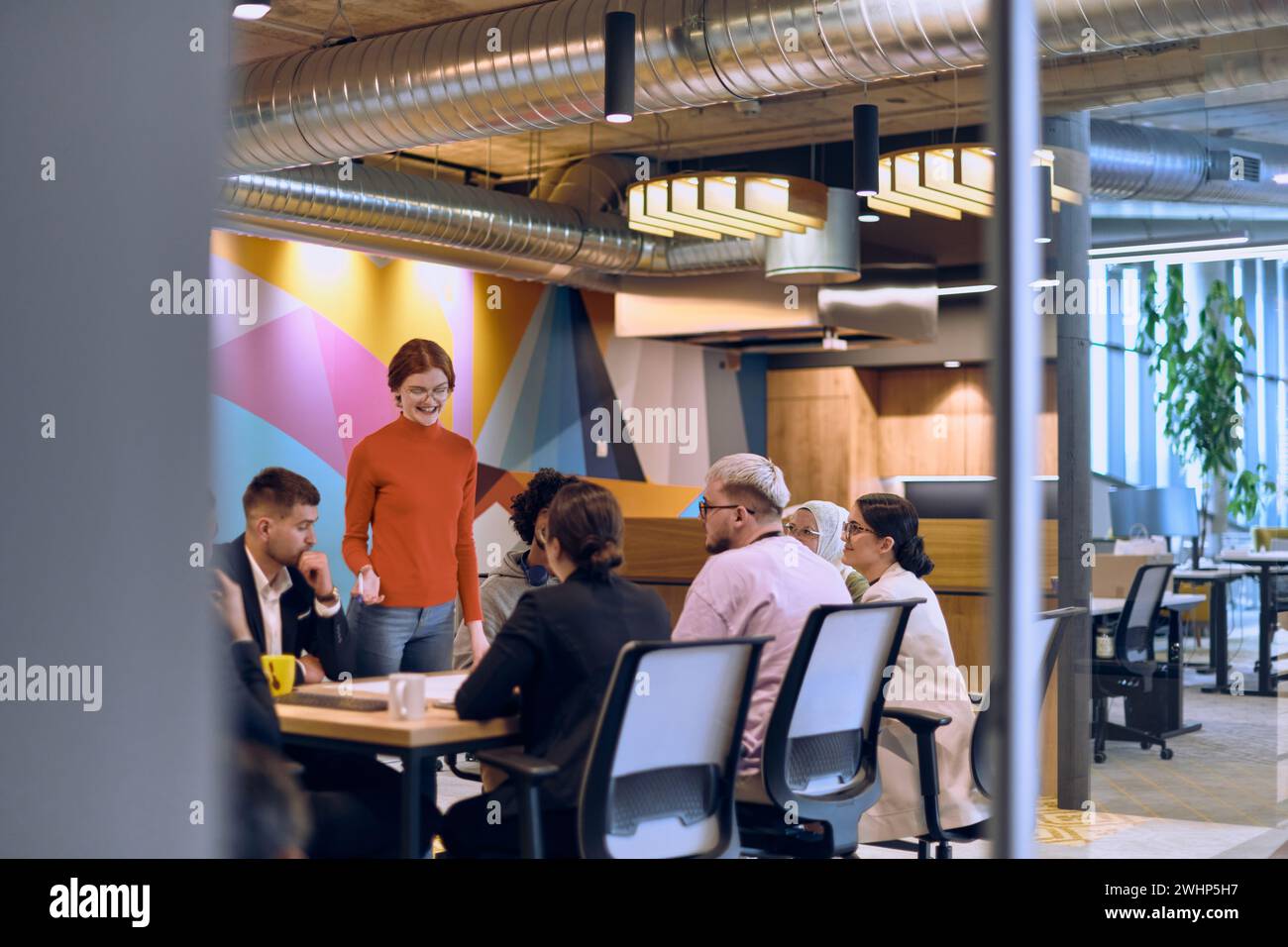 Un team diversificato di esperti aziendali in un moderno ufficio di vetro, che ascolta attentamente la presentazione di un collega, promuovendo il coll Foto Stock