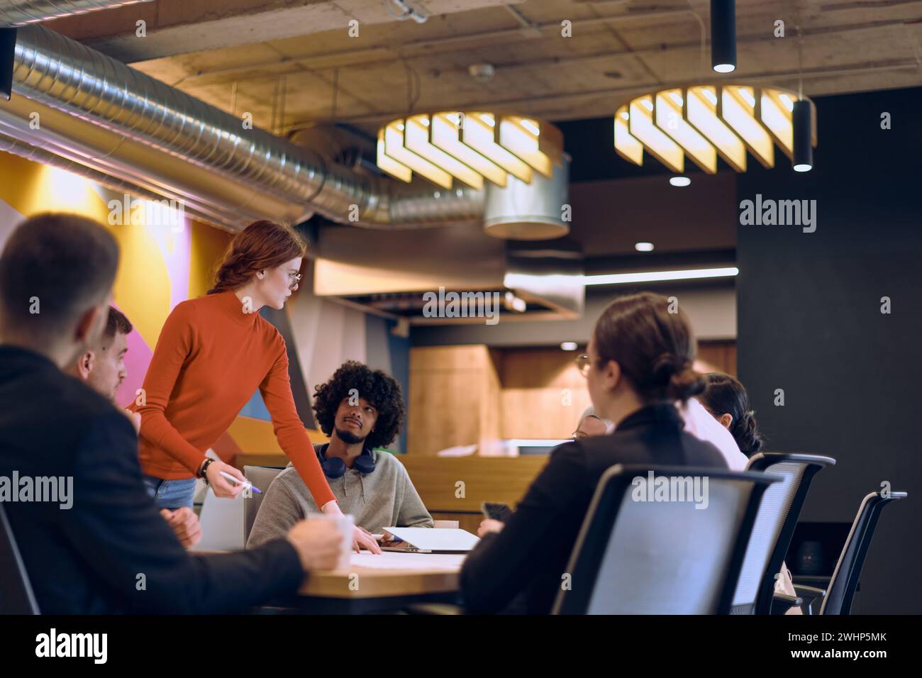 Un team diversificato di esperti aziendali in un moderno ufficio di vetro, che ascolta attentamente la presentazione di un collega, promuovendo il coll Foto Stock
