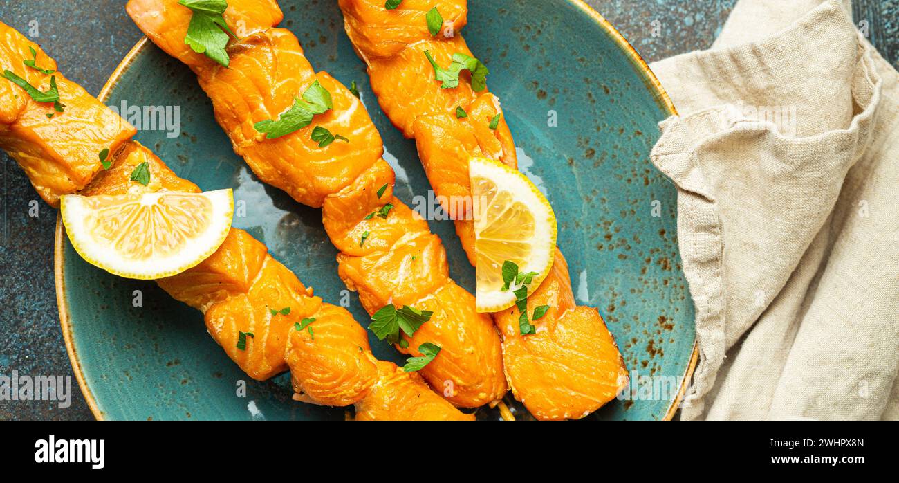 Spiedini di salmone grigliati conditi con prezzemolo verde e limone su piatto di ceramica su fondo rustico in cemento blu scuro Foto Stock