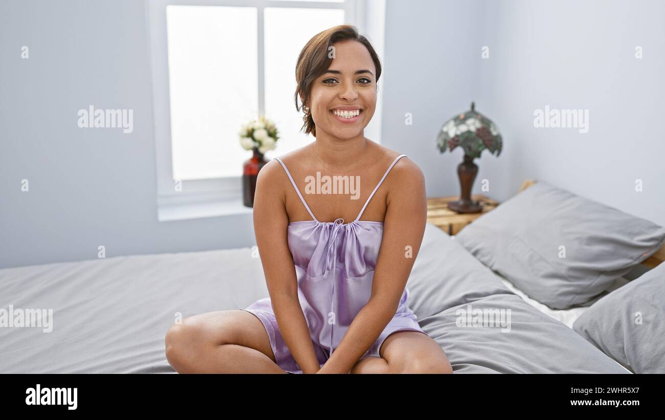 Giovane donna ispanica sorridente che si gode il relax mattutino, seduto tranquillamente sul letto, trasuda un'aura di bellezza, felicità e stile di vita positivo Foto Stock