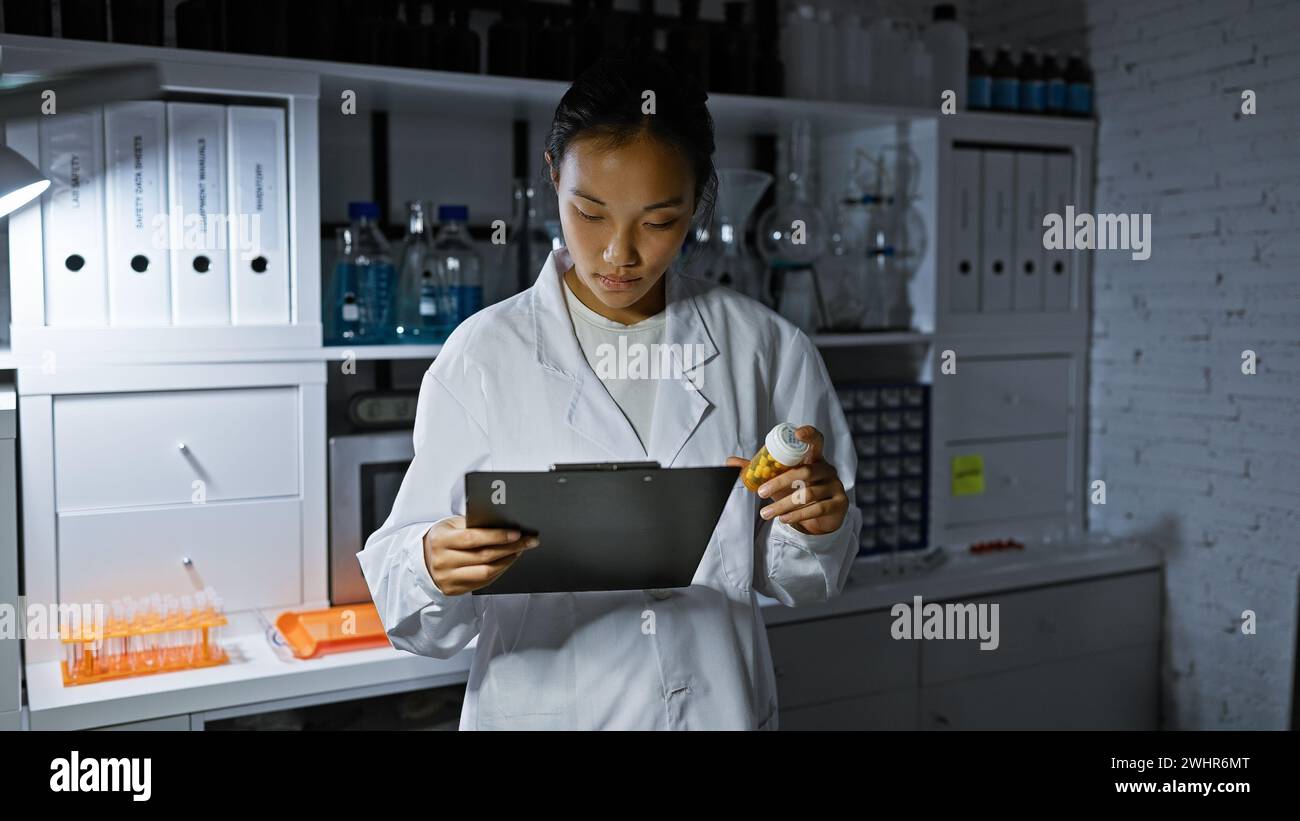 Una giovane scienziata asiatica che esamina i dati su una clipboard mentre tiene i farmaci in un ambiente di laboratorio Foto Stock