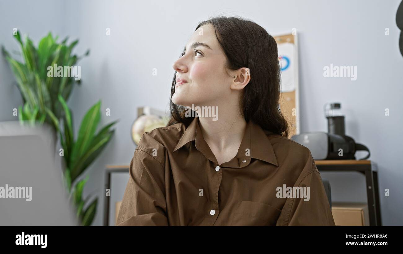 Una giovane donna con un abbigliamento casual per affari guarda verso l'alto in un moderno ambiente di ufficio con una pianta e dispositivi tecnologici sullo sfondo. Foto Stock