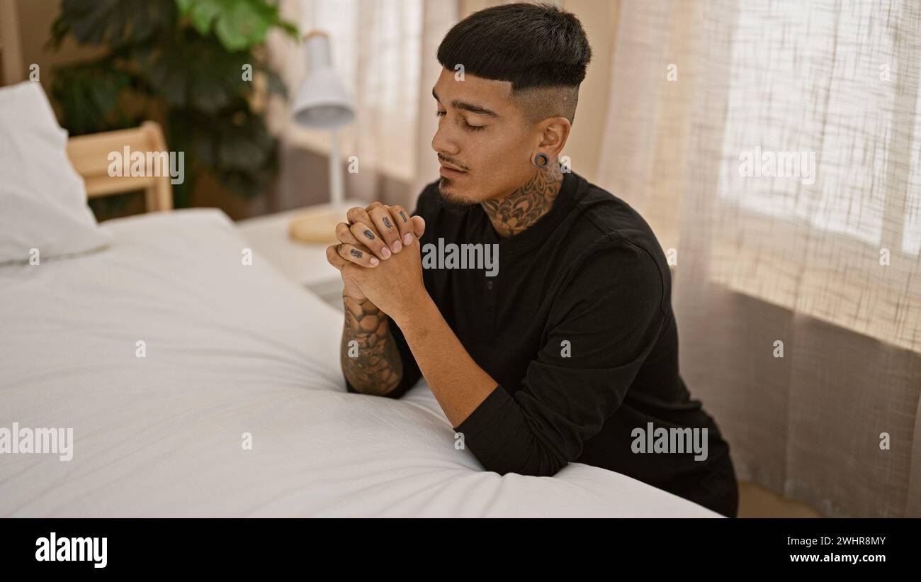 Bel giovane latino, tatuato e rilassato, seduto sul pavimento della camera da letto in pigiama, riversando la sua fede attraverso la preghiera a gesù, un ritratto di speranza Foto Stock