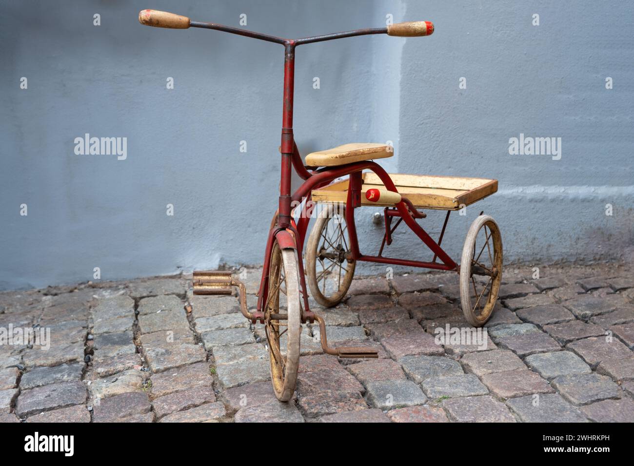 Bici vintage a tre ruote su marciapiede pavimentato in pietra. triciclo in stile retrò anni '1940 bicicletta in ferro battuto per bambini, pedale arrugginito, sella in legno, pneumatico danneggiato. Foto Stock