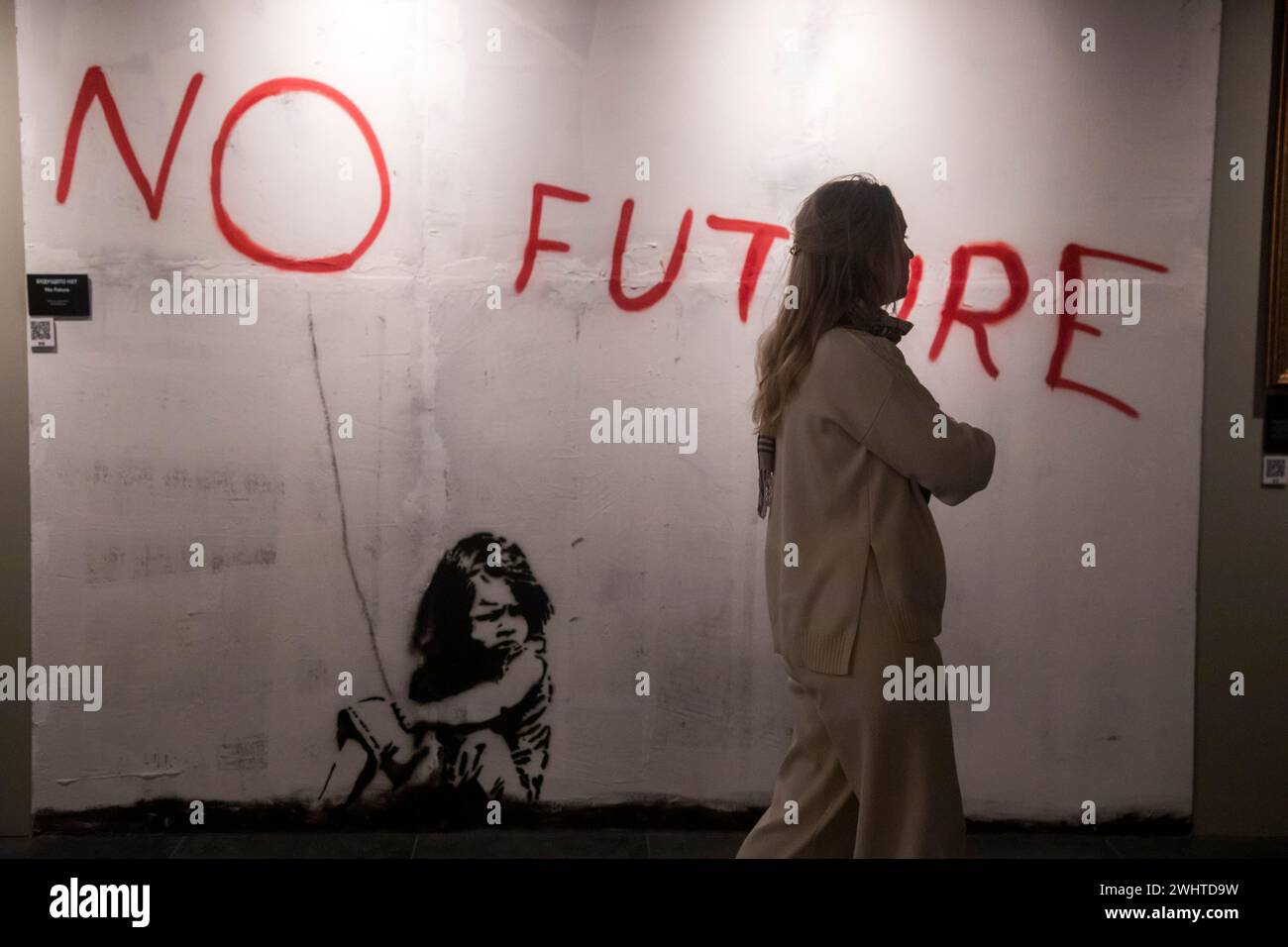 Mosca, Russia. 10 febbraio 2024. Una donna si trova davanti al dipinto "No Future" in mostra durante una mostra dedicata all'artista di strada inglese Banksy, a Mosca, in Russia. Intitolato 'Find Banksy' e tenuto al VDNKH Pavilion No 21, contiene copie delle opere originali di Banksy Foto Stock