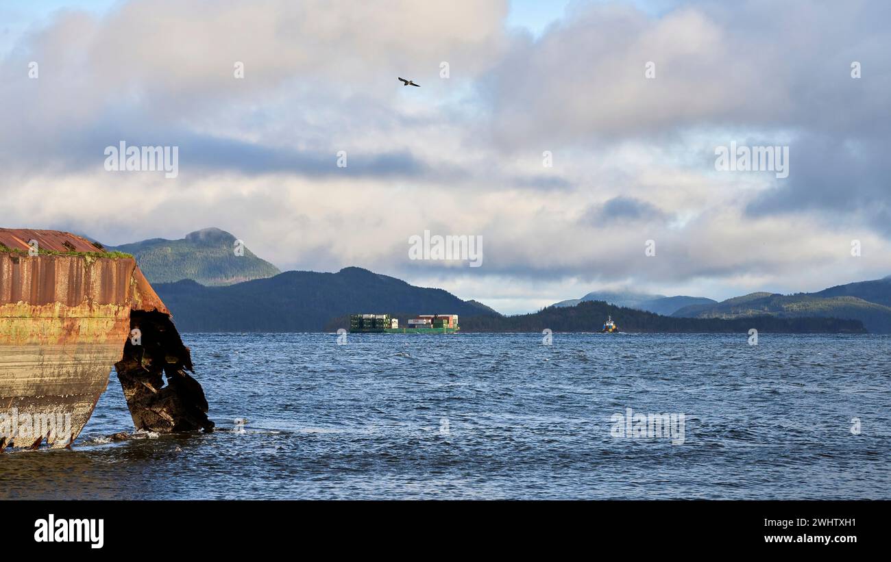Un rimorchiatore e una chiatta distanti che viaggiano lungo un'insenatura dell'oceano con lo scafo arrugginito di una vecchia nave in primo piano. Foto Stock