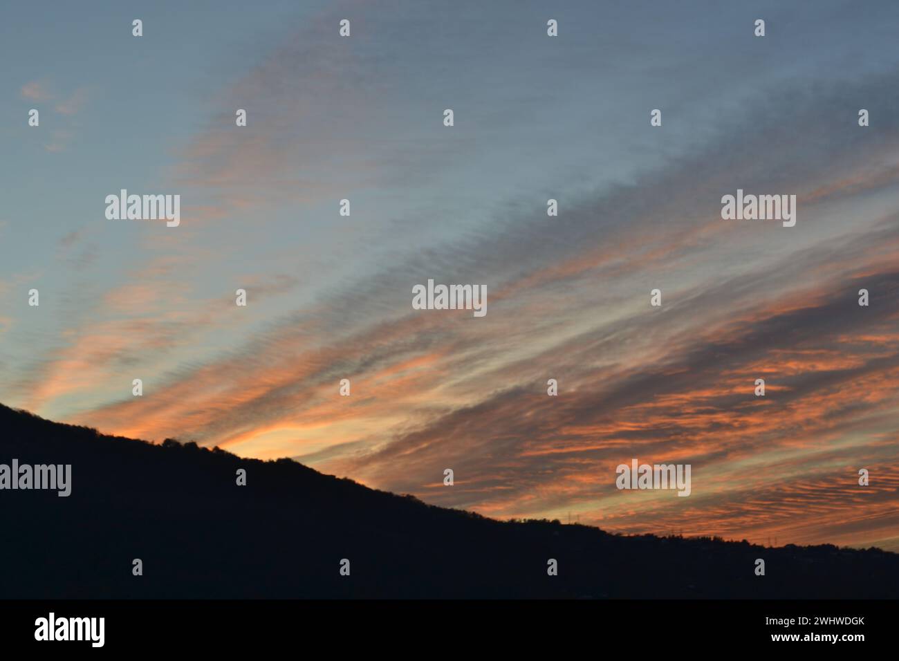 Vista Beautigul di uno strano tramonto rosso a righe e innaturalmente diretto in un senso nuvole grigie a forma di piuma sul cielo blu serale sopra la catena montuosa. Foto Stock