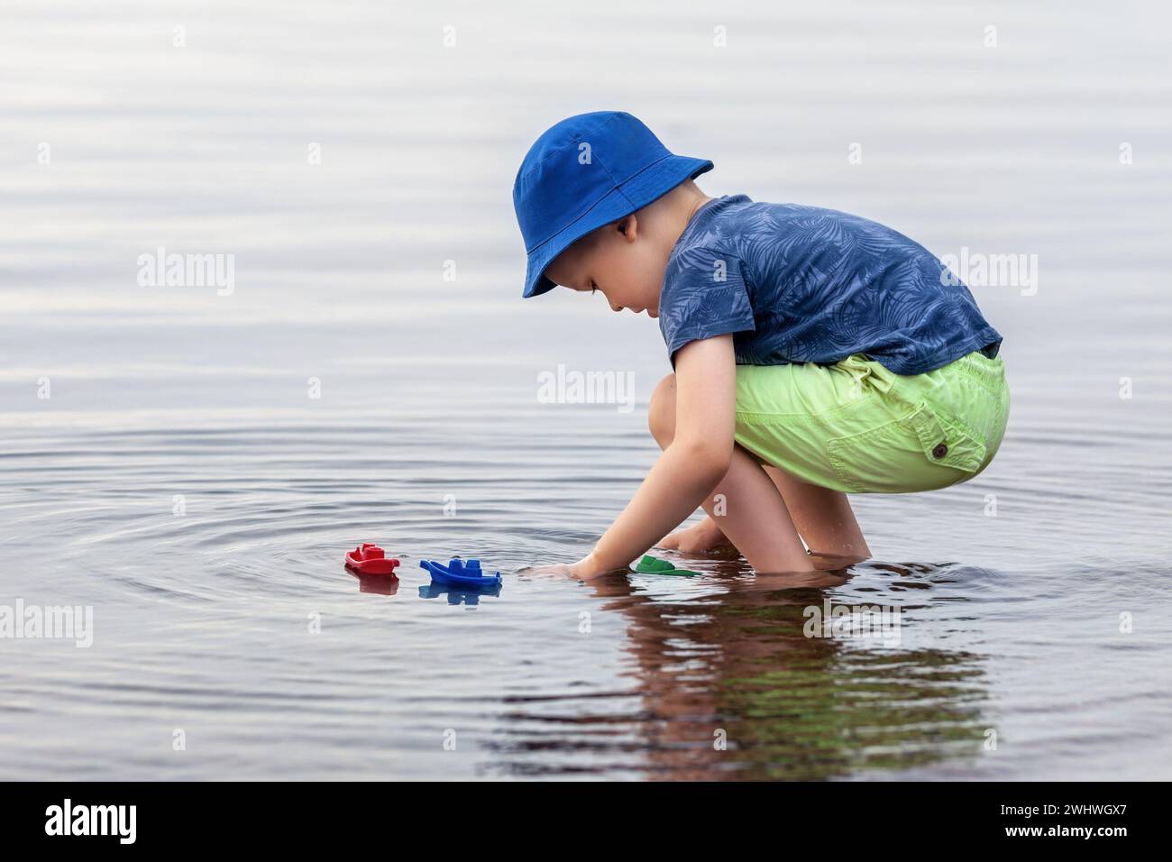 Il ragazzo si accovaccia in acque basse e calme del lago e gioca con le sue barche colorate. È ora di estate. Foto Stock