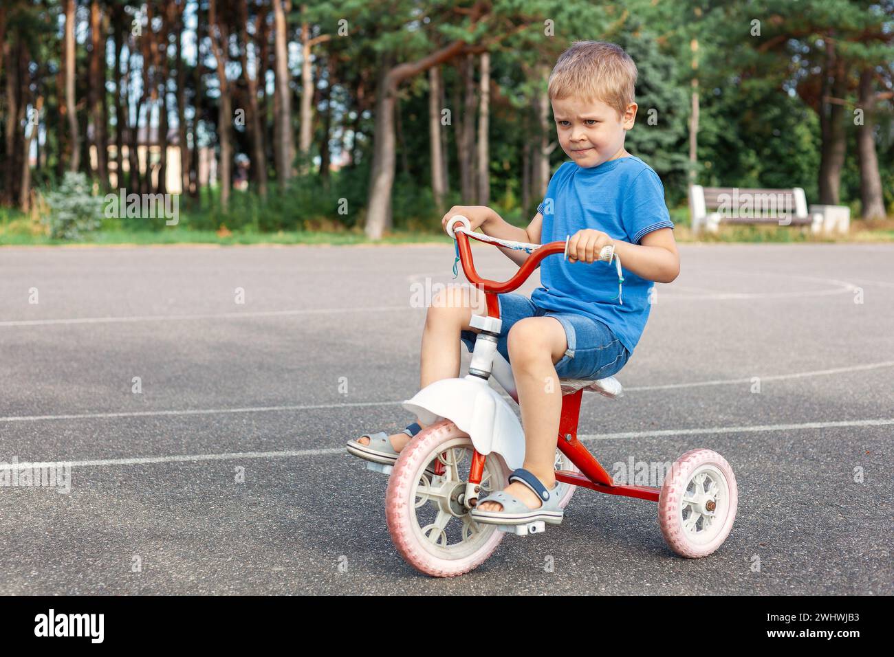 Un bambino carino molto soddisfatto sta guidando il suo vecchio triciclo nel campo da basket all'aperto della città. Foto Stock