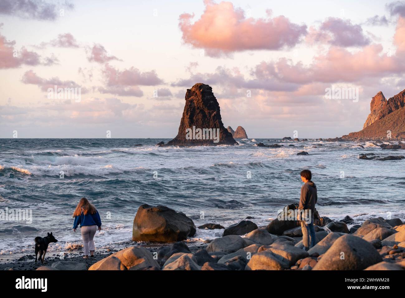 Giovani coppie felici che si godono un tramonto sulla spiaggia con il loro cane. Spiaggia di Benijo a Tenerife. Foto di alta qualità Foto Stock