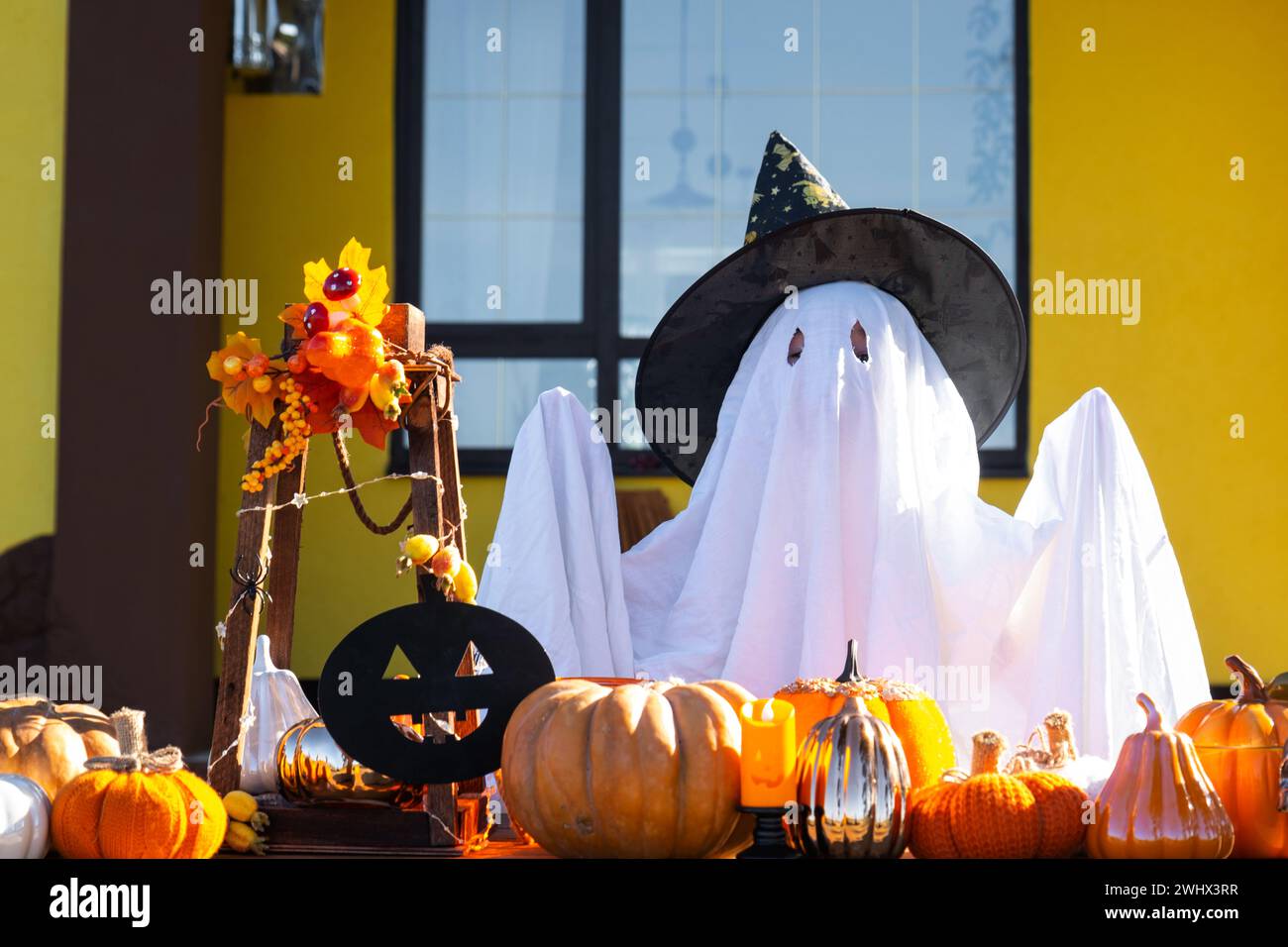 Bambino in lenzuola con fessure come fantasma nel cappello da strega e decorazioni di Halloween sul portico della casa all'esterno nel cortile della puma Foto Stock