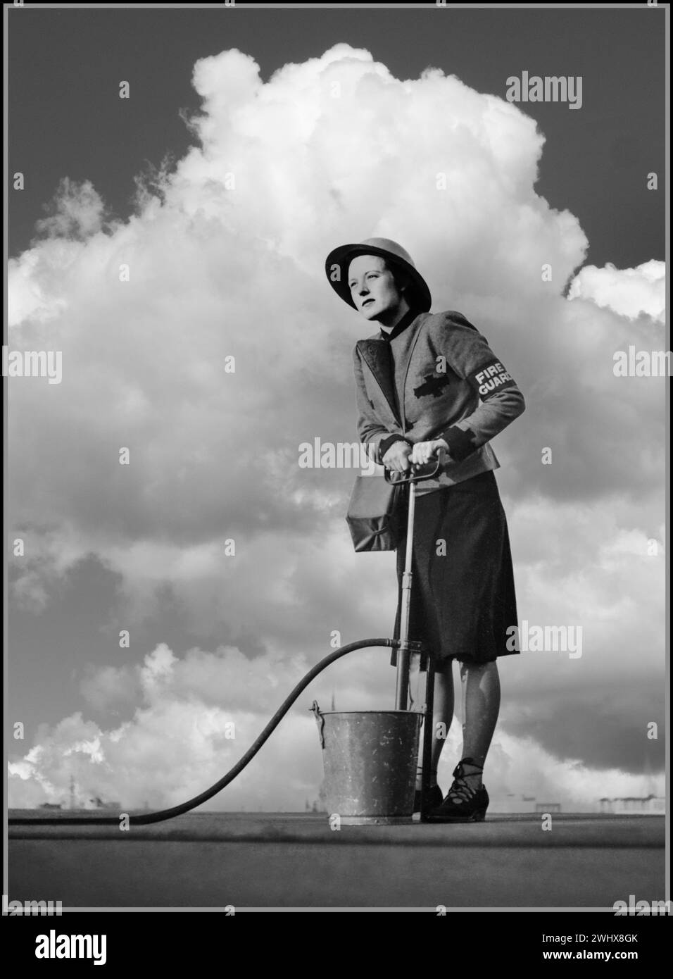 1941 donna della seconda guerra mondiale guardia del fuoco in uniforme immagine Propaganda donne in guerra Gran Bretagna Regno Unito che utilizzano una pompa a staffa in caso di incendio, sul tetto di un edificio a LONDRA Regno Unito seconda guerra mondiale Una guardia del fuoco donna che dimostra una pompa a staffa sul tetto di un edificio di Londra, indossa la fascia da braccio della Guardia antincendio, un casco d'acciaio e porta con sé la maschera antigas. Foto Stock