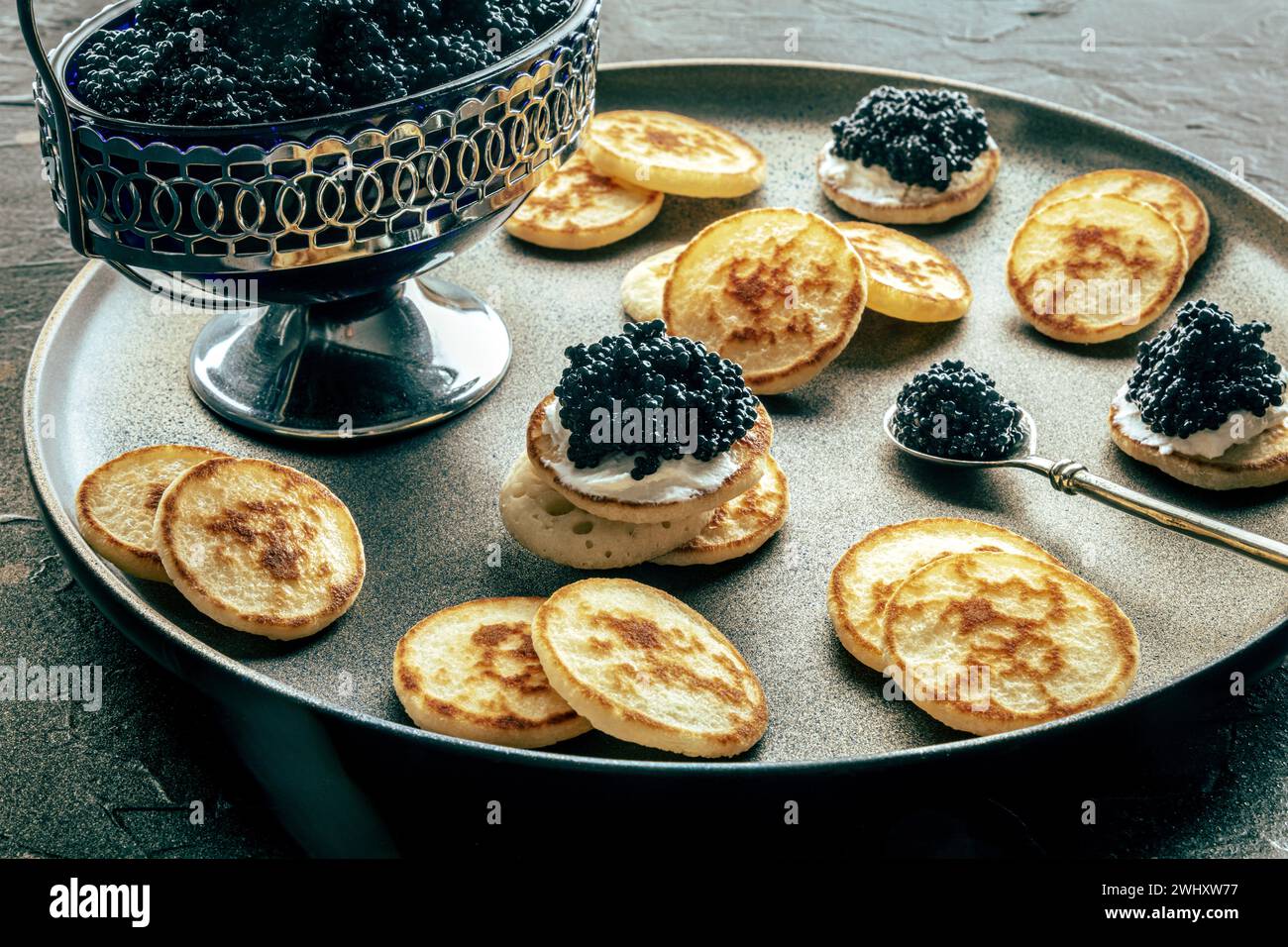 Blinis con caviale nero, su un piatto festivo su sfondo nero, mini pancake Foto Stock