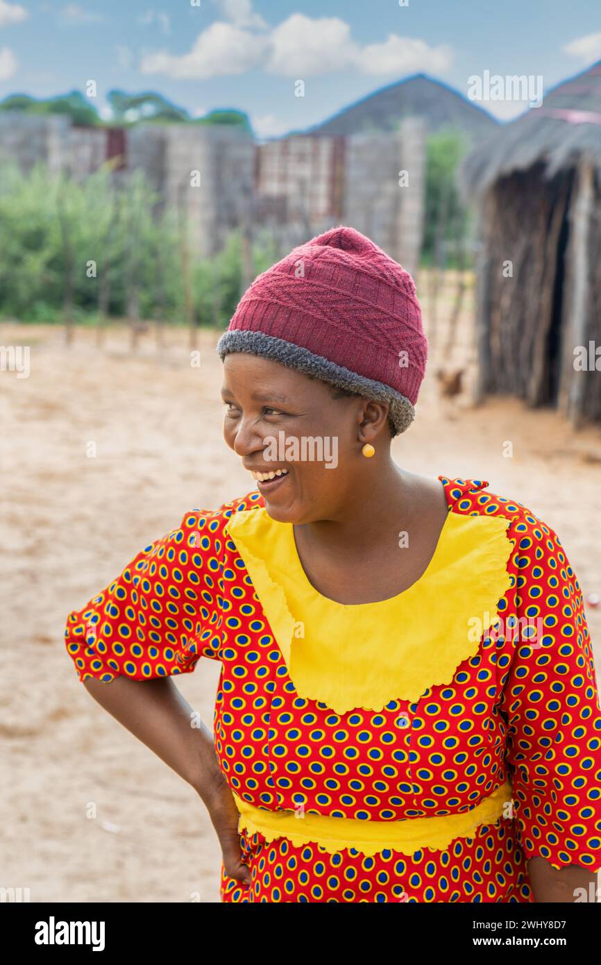 donna africana felice nel villaggio che indossa abiti tradizionali, capanna sullo sfondo nel cortile Foto Stock