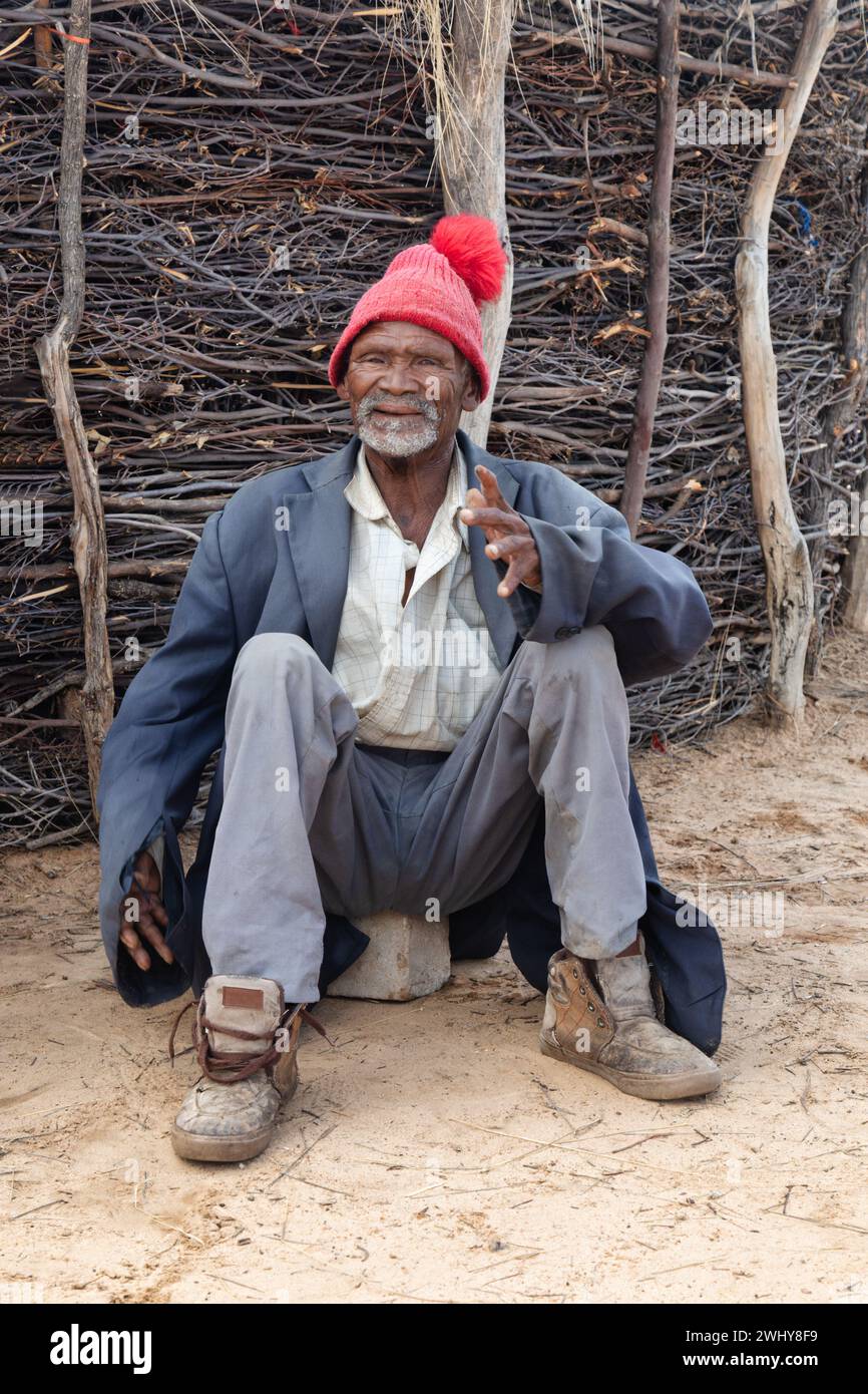 vecchio uomo africano sorridente nel cortile che si riposa, di fronte a una tradizionale recinzione kraal, vecchio abbigliamento casual e un berretto rosso Foto Stock