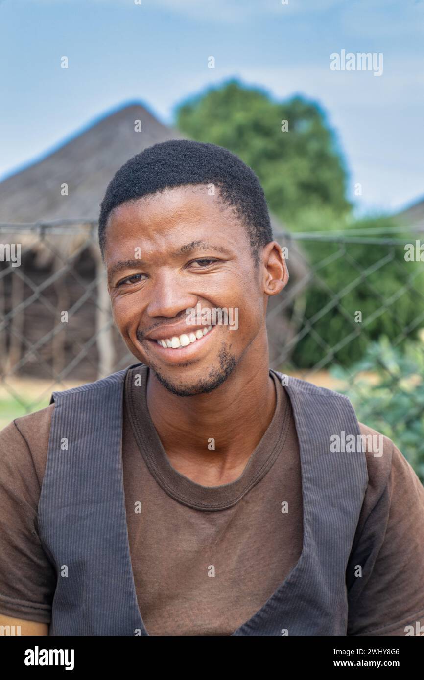 l'assistente sociale volontario del villaggio, giovane africano sorridente con un giubbotto nel cortile che si riposa, capanna tradizionale e rondavel sullo sfondo Foto Stock