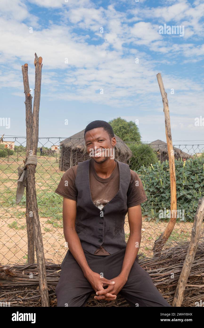 giovane villaggio africano sorridente con un gilet posto sulla recinzione nel cortile, capanna tradizionale e rondavel sullo sfondo Foto Stock