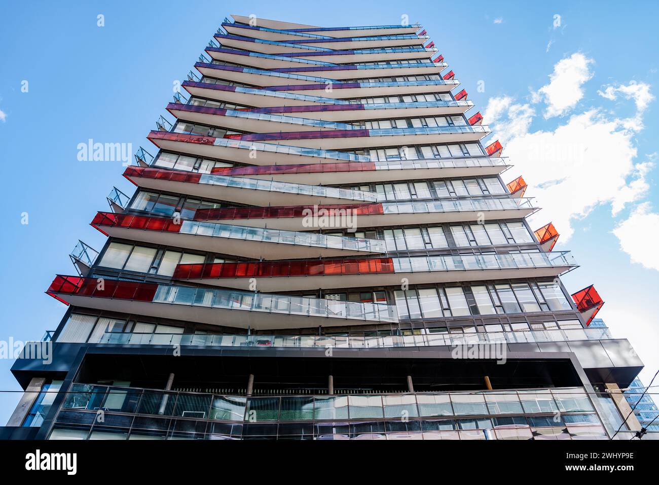 Un moderno edificio a piu' piani con balconi sfalsati che presentano vibranti accenti rossi, adagiato su un cielo limpido con nuvole volose Foto Stock