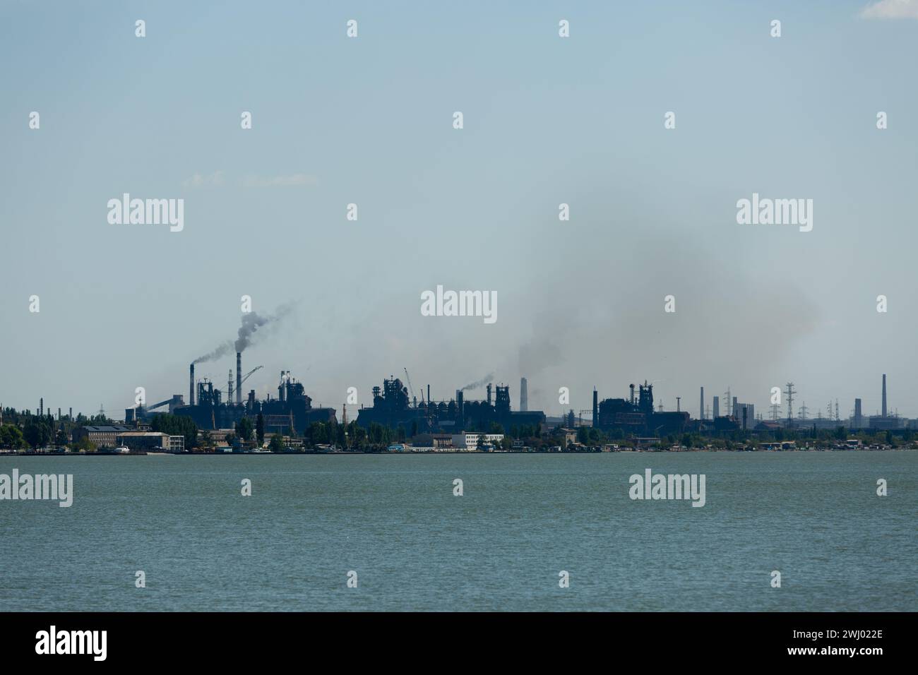Skyline industriale dominato dalle emissioni degli impianti metallurgici, fumo che si disperde contro il cielo, simboleggia i problemi di inquinamento ambientale, con l'acqua in primo piano. Foto Stock