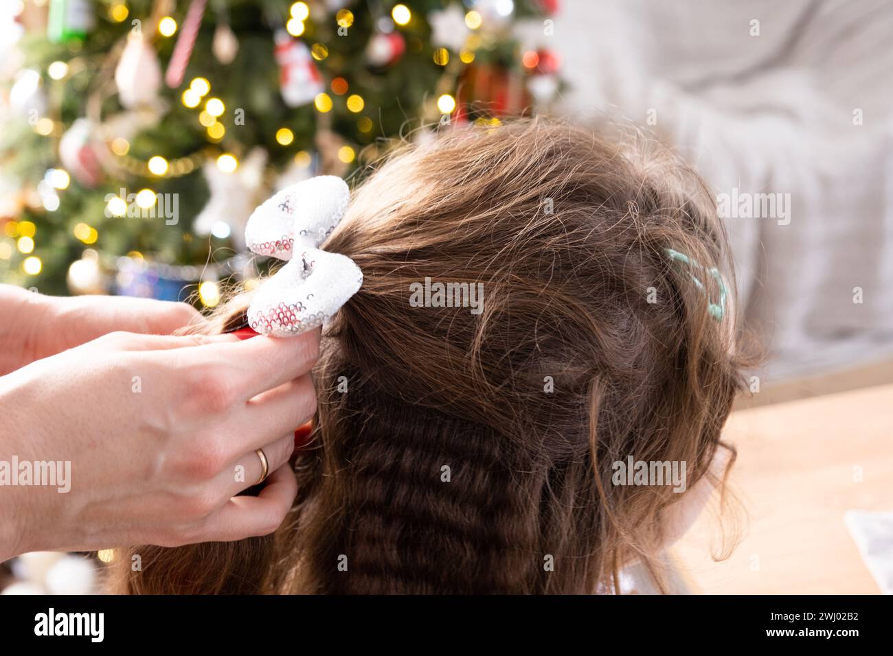 Capelli lunghi di una ragazza con un fiocco lucente - le mani della mamma pettinano la figlia per una vacanza contro le luci dell'albero di Natale Foto Stock