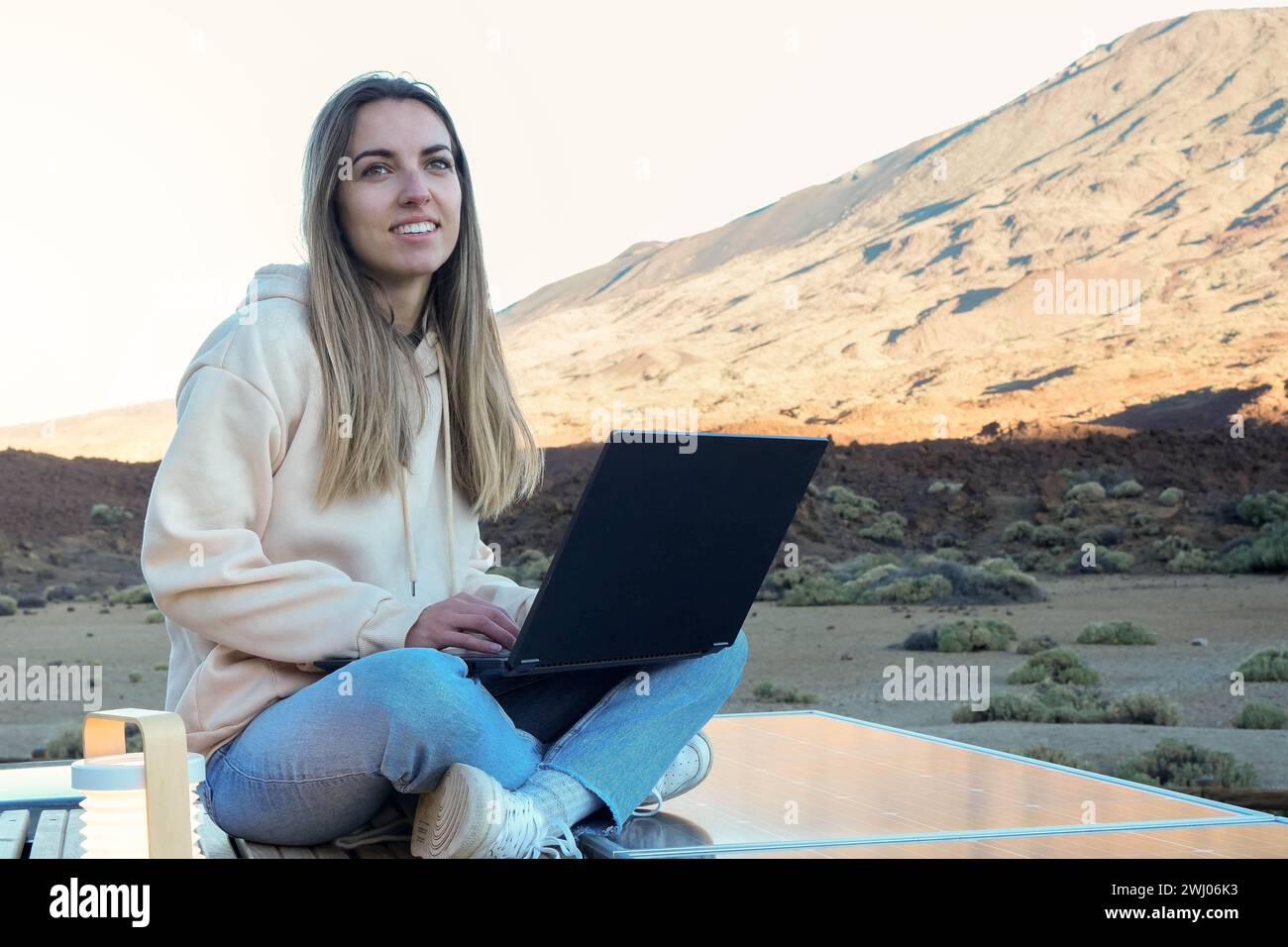 Una giovane donna viene vista sedersi sul tetto di un furgone, utilizzando un computer portatile in un pittoresco ambiente esterno. Foto Stock