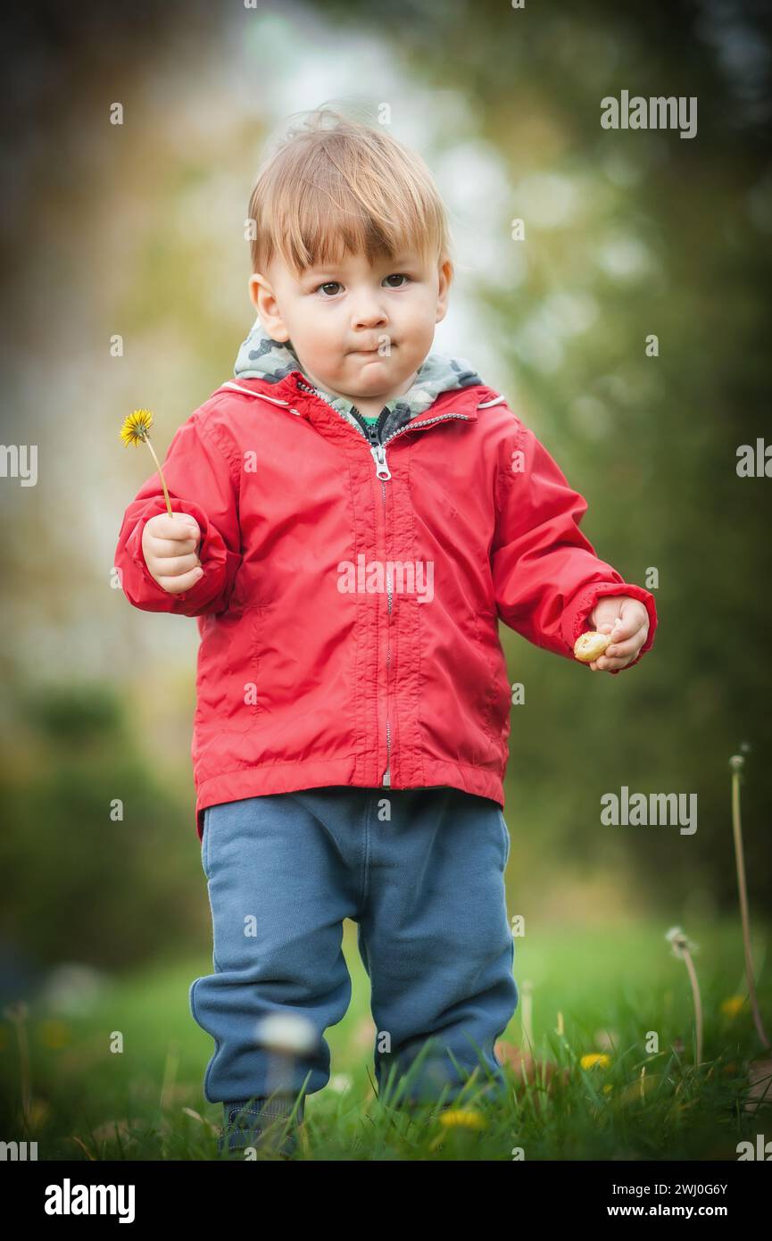 Il grazioso bambino, adornato da una vivace giacca rossa, scopre l'incanto della natura mentre culla con affetto un delicato fiore Foto Stock