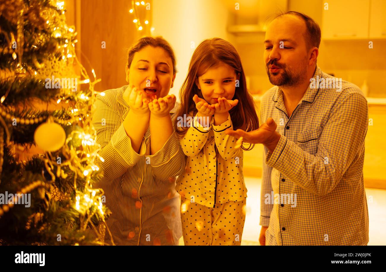 Una bambina carina con i suoi genitori farà saltare via i coriandoli natalizi. Marito e moglie con la figlia in pigiama vicino al Chri Foto Stock